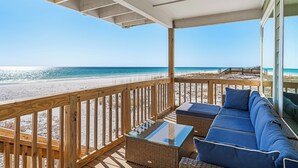 Un balcon donnant sur la plage, avec un canapé en osier et une table à plateau en verre, d’où l’on peut admirer une plage de sable et l’océan.