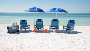 Une scène de plage avec quatre chaises de plage à rayures et deux parasols bleus, une glacière Coleman et un ballon de football posé sur l’une des chaises.