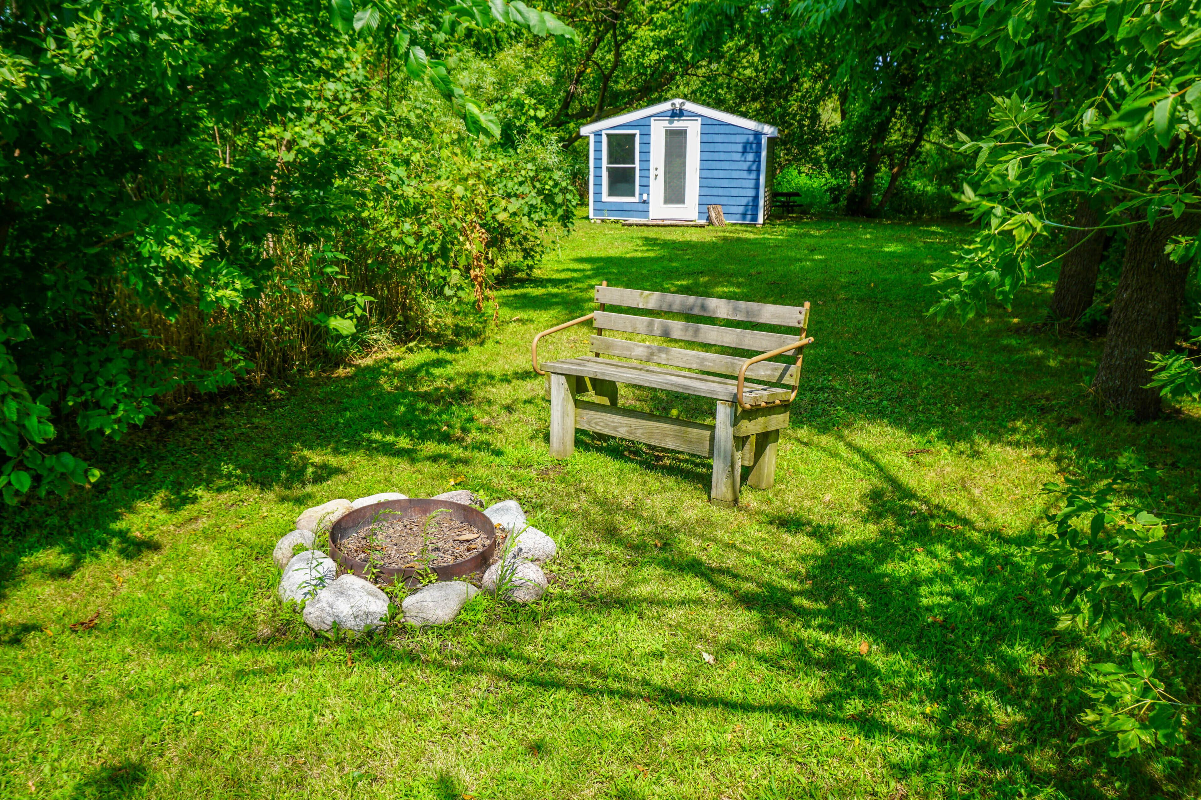 A wooden bench in a grassy area with a small fire pit and a blue shed in the background.