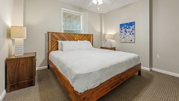 A bedroom with a wooden bed, two bedside tables, a ceiling fan, a window with blinds, and a wall art piece.