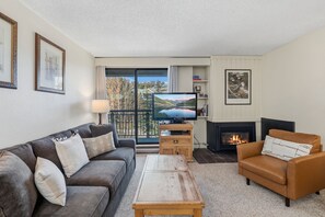 A living room with a grey sofa, a brown armchair, a wooden coffee table, a flat-screen TV, a fireplace, and a balcony with a scenic view.