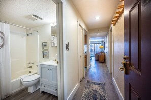 A hallway with a bathroom on the left, featuring a toilet, sink, and shower. The hallway leads to a living area with a sofa and a lamp.