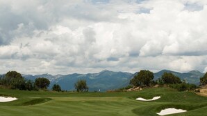 Un campo de golf con búnkeres de arena, una bandera y montañas al fondo.