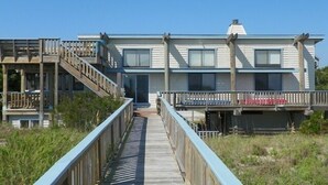 A two-story beachfront house with a wooden deck leading to the entrance, a covered porch, and a small staircase to the upper level. - WH1091- Oceanfront-  6 BRs, Cargo Lift, Great Ocean Views Dog Friendly! (Corolla)