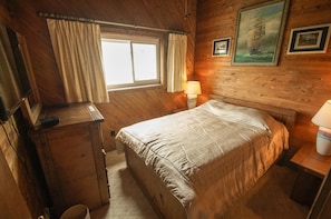 Quarto de cabana de madeira com cama, escrivaninha, janela com cortinas e abajur.