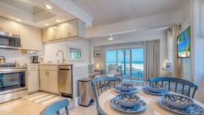 A modern kitchen with built-in appliances, a dining area with blue chairs and plates, and a view of the beach through large windows.
