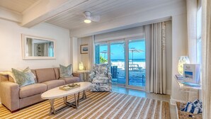A spacious living room with a beige sofa, a coffee table, and a patterned rug. Large windows offer a view of the beach.