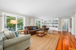 A spacious living room with a large window, a bookshelf, and a wooden floor. - Long Pond Escape—Decks, Kayaks & Fishing (Harwich)
