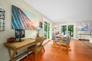 A dining area with a wooden table and chairs, a painting on the wall, and a view through the glass door. - Long Pond Escape—Decks, Kayaks & Fishing (Harwich)