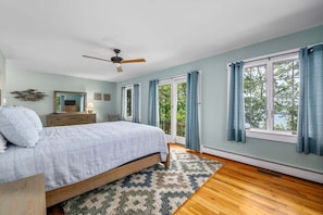 A bedroom with a bed, a dresser, a ceiling fan, a window with curtains, and a view of trees. - Long Pond Escape—Decks, Kayaks & Fishing (Harwich)