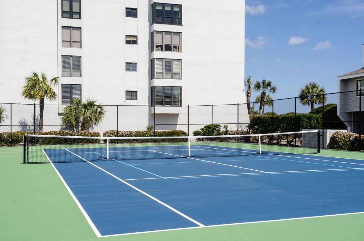 Een tennisbaan met een net, omringd door een hek, palmbomen en een gebouw op de achtergrond.