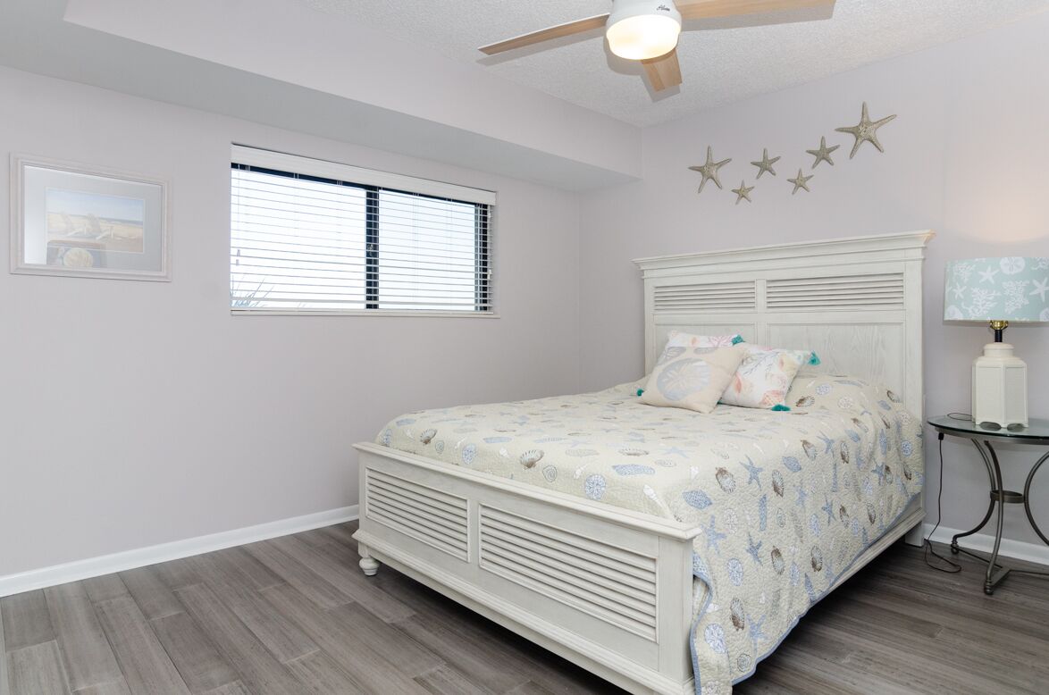 A bedroom with a bed, a ceiling fan, a window with blinds, a bedside table with a lamp, and star decorations on the wall.