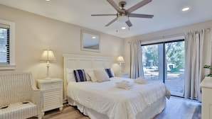 A bedroom with a large bed, two bedside tables with lamps, a ceiling fan, a window with curtains, and a wicker chair. - 2311 Racquet Club: 3BR 2 BA Villa. Community Pool and Clay Tennis, Free Bikes (Hilton Head Island)