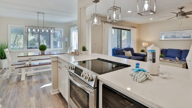 A modern kitchen with a central island, built-in oven, and stainless steel appliances. There is a dining area with a white table and chairs, and a living area with a blue sofa and a ceiling fan. - 2311 Racquet Club: 3BR 2 BA Villa. Community Pool and Clay Tennis, Free Bikes (Hilton Head Island)