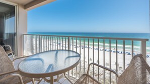 A balcony with a glass table and chairs, overlooking a beach with umbrellas and people.
