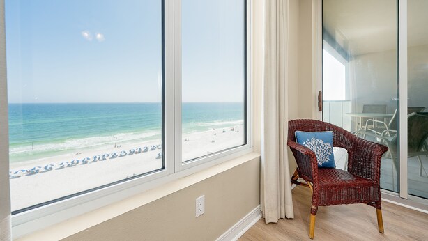 A room with a large window overlooking a beach, a wicker chair with a blue pillow, and a sliding glass door.