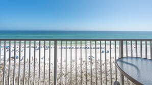 A beach view from a balcony with a table and chairs, beach umbrellas, and sand.