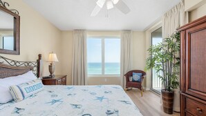 A bedroom with a bed, a nightstand, a lamp, a chair, a large window, and a plant.