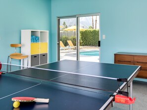 Uma mesa de tênis com rede, uma cadeira, um armário pequeno e vista para uma piscina e montanhas através de uma porta de vidro.