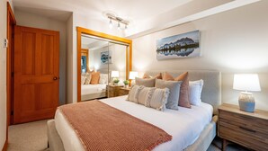 Een slaapkamer met een groot bed, een houten deur, een spiegel, een nachtkastje met een lamp en een schilderij aan de muur.