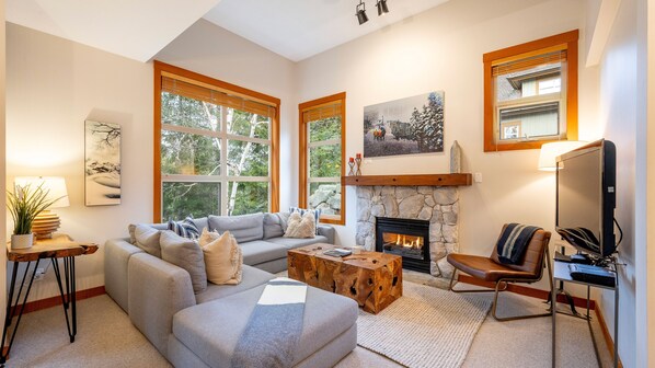 A cozy living room with a fireplace, large windows, a sofa, a coffee table, and a TV.