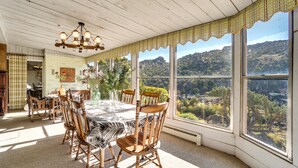 Uma sala de jantar com mesa posta para seis pessoas, rodeada por cadeiras de madeira, janelas amplas com vista panorâmica e um lustre.