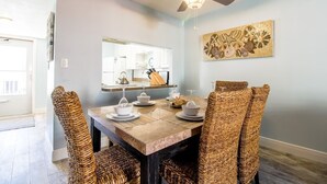 Un comedor con una mesa preparada para cuatro personas, sillas de mimbre, un ventilador de techo y una cocina visible a través de una puerta abierta.