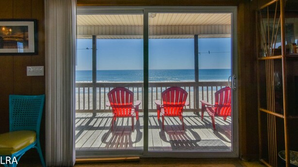 A room with a view of the ocean, featuring a sliding glass door, two red Adirondack chairs, and a wooden deck. - The Choice: Oceanfront Comfort in Surf City (Surf City)