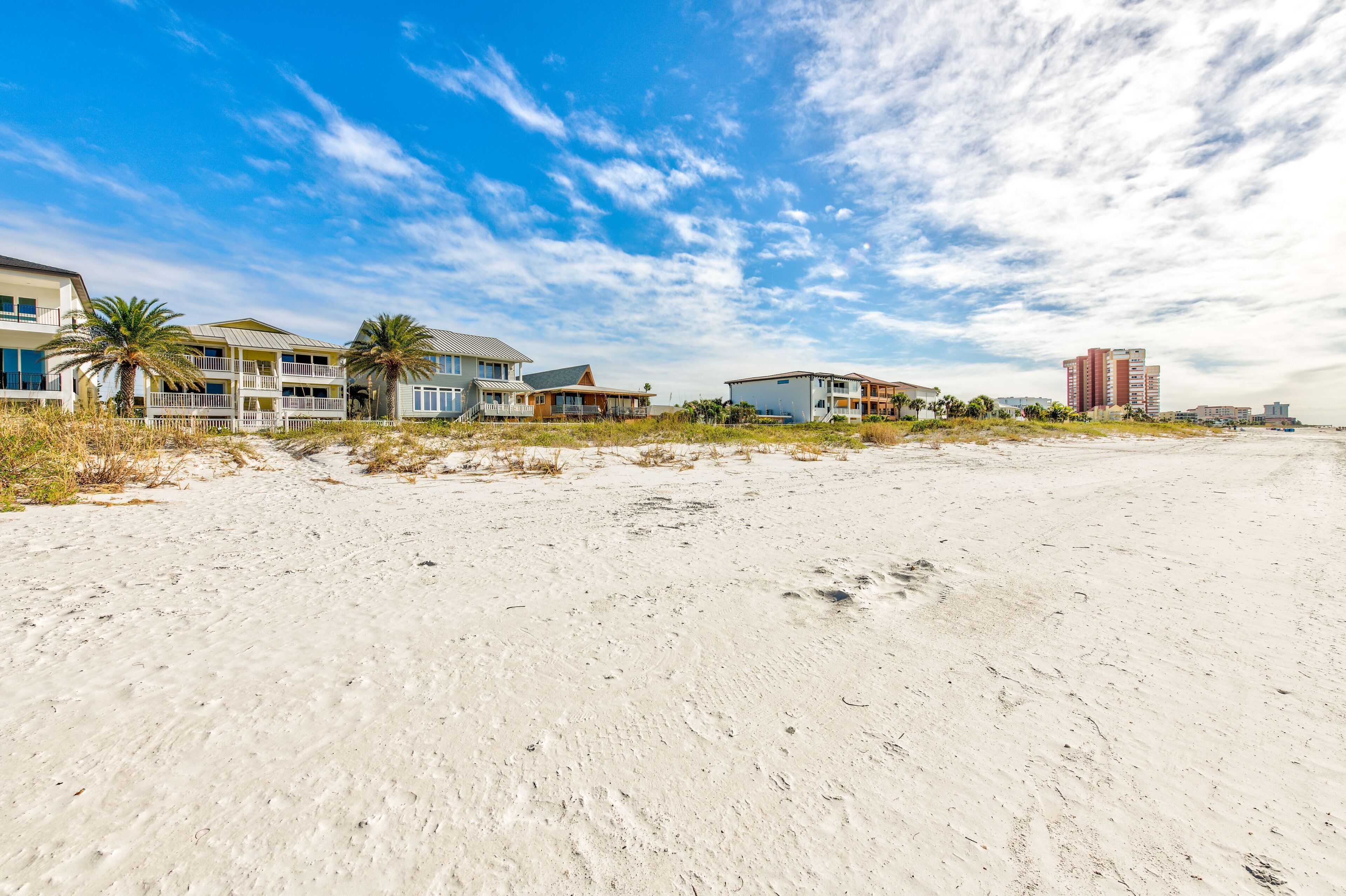 A beachfront property with multiple buildings, including a large hotel and residential houses, with a sandy beach and palm trees in the foreground.