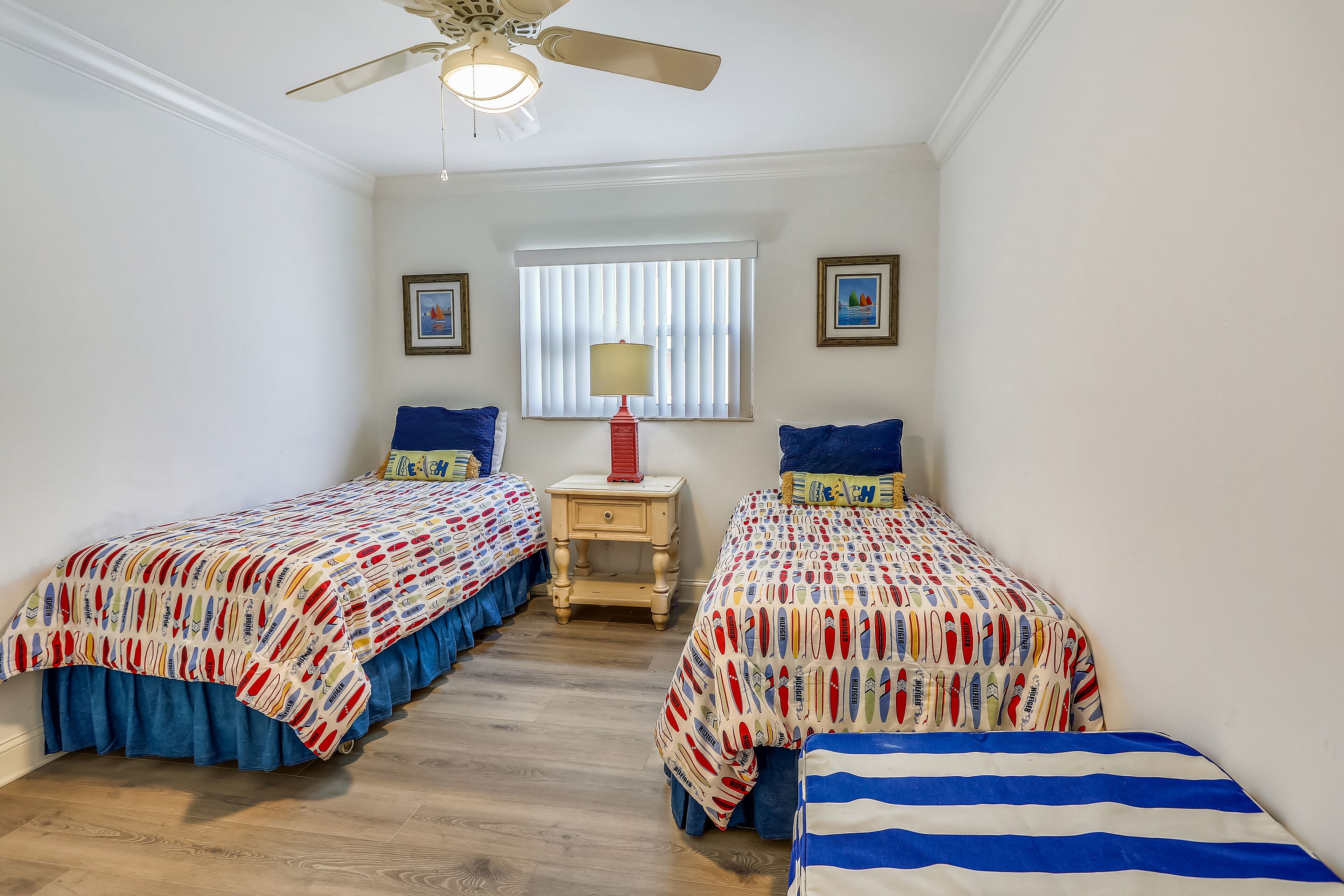 Een slaapkamer met twee bedden, een plafondventilator, een raam met jaloezieën en een nachtkastje met een lamp.