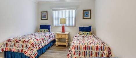 Un dormitorio con dos camas, un ventilador de techo, una ventana con persianas y una mesita de noche con lámpara.