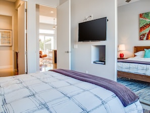 Quarto de hotel com cama, televisão na parede, um prateleira pequena e uma janela.