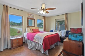 A bedroom with a bed, a nightstand, a dresser, a ceiling fan, and large windows with curtains. - 3 Bedroom/3 Bath home overlooking 2 Creeks with Hot Tub and Fire Pit! (Wimberley)