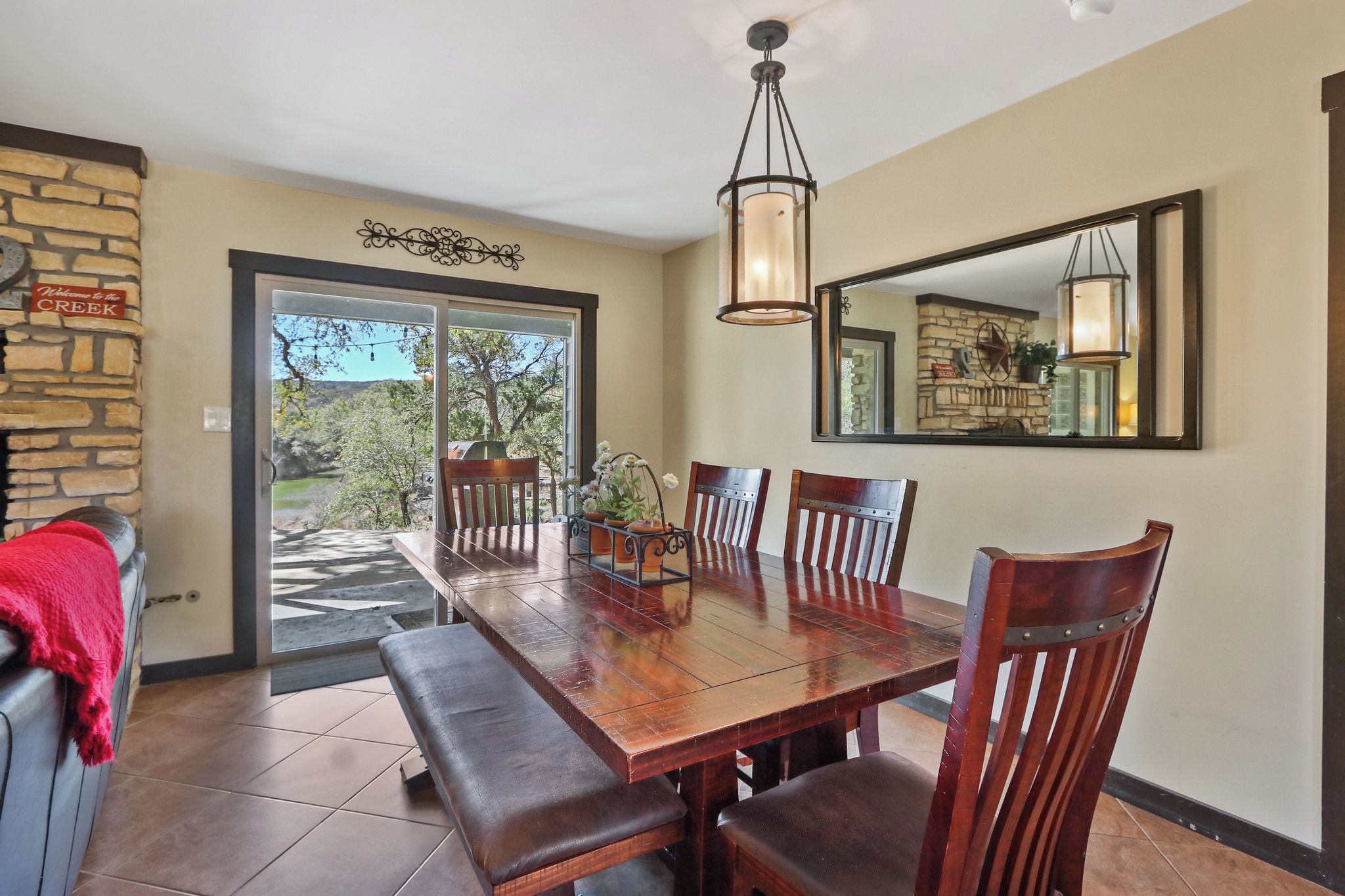 A dining area with a wooden table, benches, and chairs. There is a stone wall with a sign, a large mirror, and a hanging light fixture.