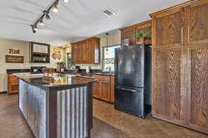 A modern kitchen with wooden cabinets, a black refrigerator, and a central island. - 3 Bedroom/3 Bath home overlooking 2 Creeks with Hot Tub and Fire Pit! (Wimberley)