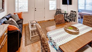 Ein Wohnzimmer mit einer Couch, einem Couchtisch und Stühlen, einem Esstisch mit Platzdeckchen, einem Fernseher und einer Tür mit Fenster.