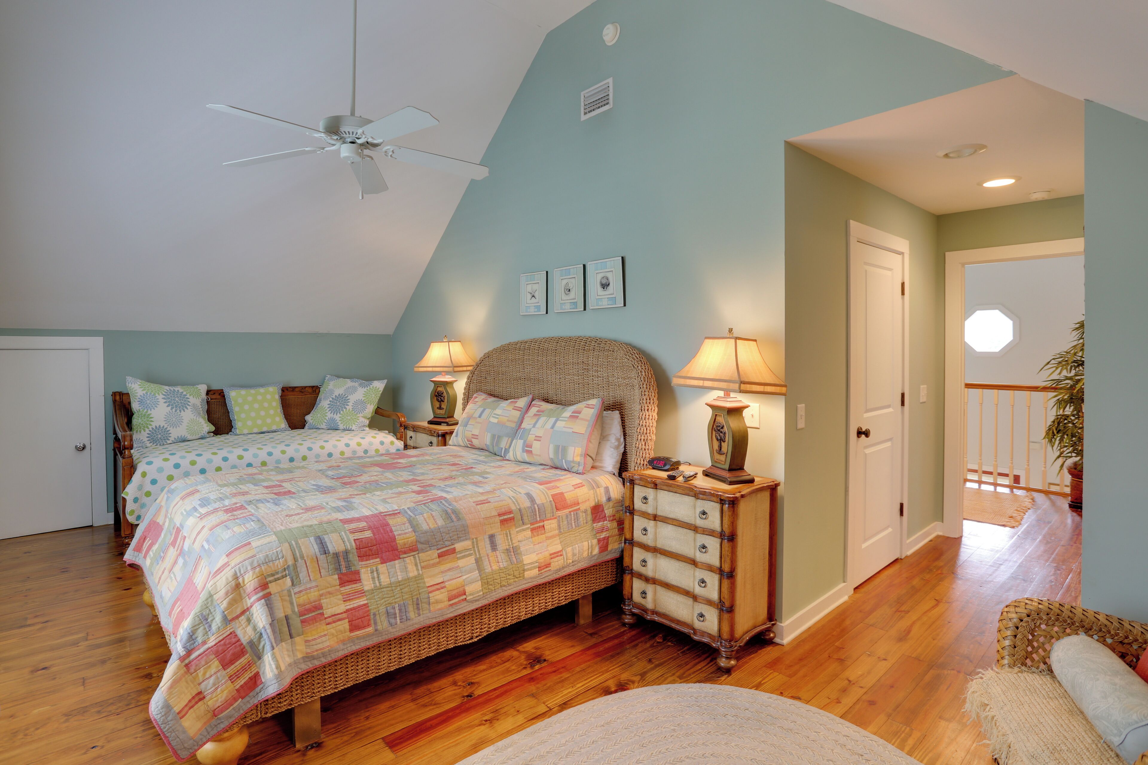A bedroom with a large bed, a ceiling fan, a nightstand with a lamp, and a door leading to another room.