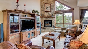 A spacious living room with a stone fireplace, a large flat-screen TV, leather furniture, and large windows offering a view of the outdoors. - Private Hot Tub, Ski-in/ski-out! (Breckenridge)