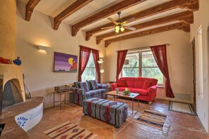 A cozy living room with a stone fireplace, wooden beams, a red sofa, patterned ottoman, and a stone floor.