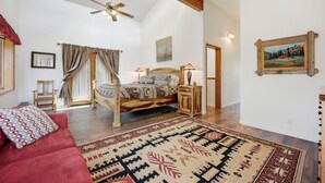Une chambre spacieuse avec un grand lit, un ventilateur de plafond, une tête de lit en bois, une table de chevet, un canapé rouge et un tapis à motifs.