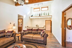 A spacious living area with a large brown leather sofa, a wooden coffee table, and a lamp. There is a wooden staircase leading to an upper level with windows and a shelf with decorative items. - Large Home on 1 Acre: 2 Mi to Grand Lake Village (Grand Lake)