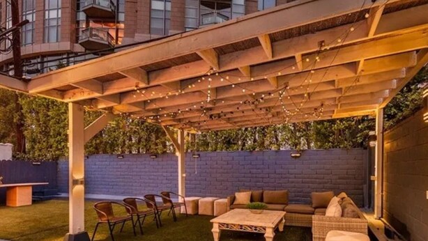 A covered outdoor patio with a seating area, including a sofa and chairs, under a wooden pergola with string lights. - Luxurious 3-Bedroom Westwood Gem  (Los Angeles)