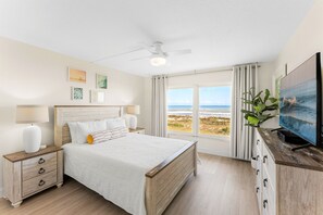 A bedroom with a bed, nightstands, a TV, and a view of the beach.