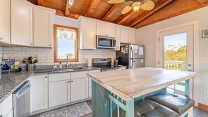 A kitchen with white cabinets, a microwave, a stove, and a refrigerator. There is a window with a view of trees and a balcony with a railing. - Duck Life - Oceanside, Sound View, 325 Yards to Beach, Pool, Hot Tub, Tiki Bar, Pool Table, Pets OK (Duck)