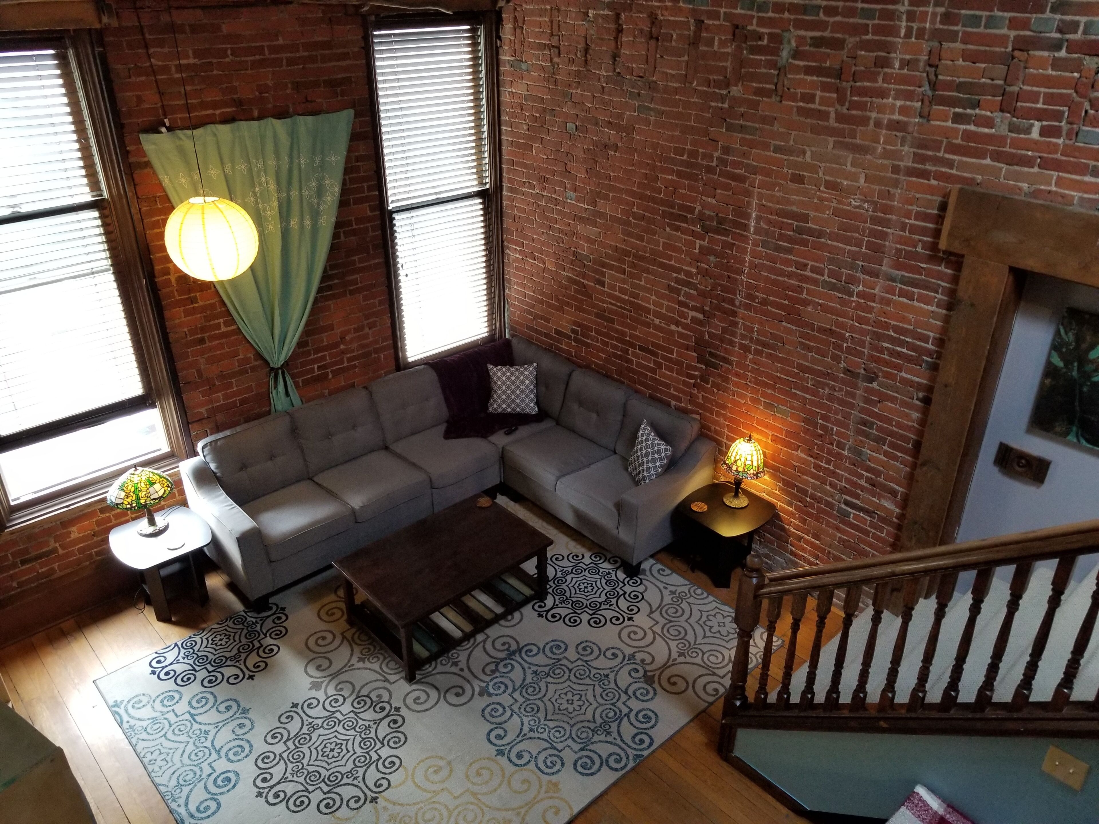 Ein Loft-Wohnzimmer mit freiliegenden Backsteinwänden, einem grauen Ecksofa, einem Holztisch, gemusterten Teppichen und einer Treppe mit Holzwand.