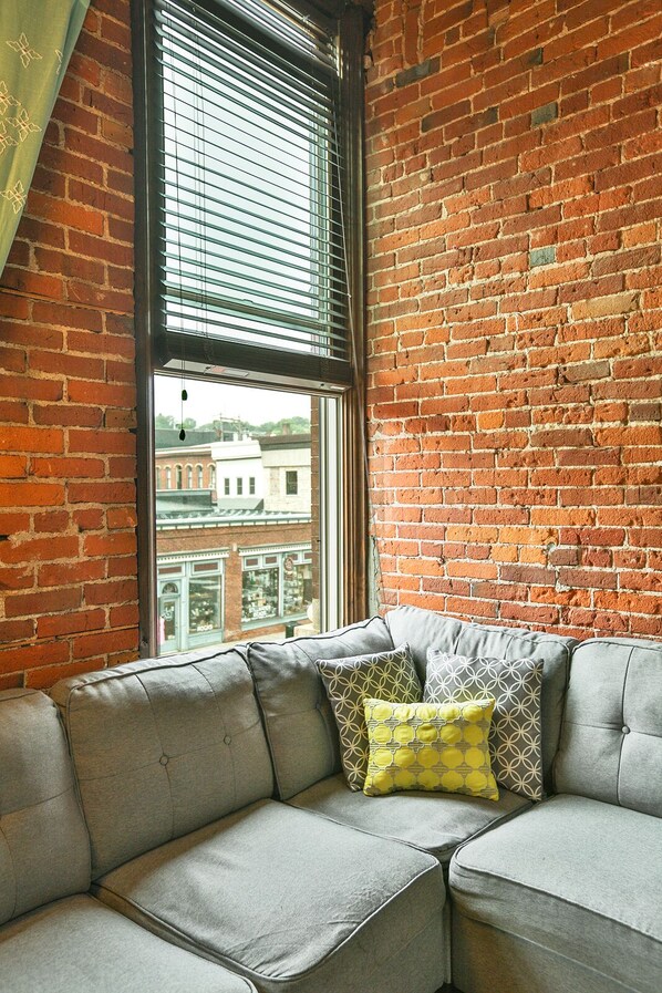 A room with a brick wall, a large window with blinds, and a grey upholstered sofa with patterned cushions.