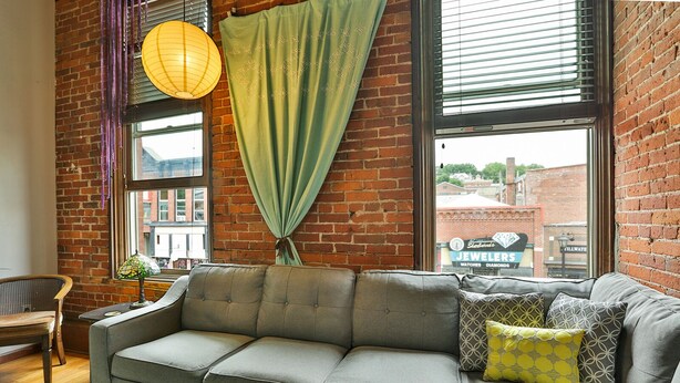 A cozy living room with a grey sofa, patterned cushions, a hanging lamp, and a view of a brick building outside.