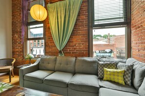 A cozy living room with a grey sofa, patterned cushions, a hanging lamp, and a view of a brick building outside.