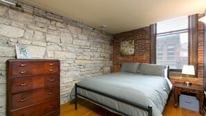 A bedroom with a stone wall, a wooden dresser, a metal bed frame, and a window with blinds.