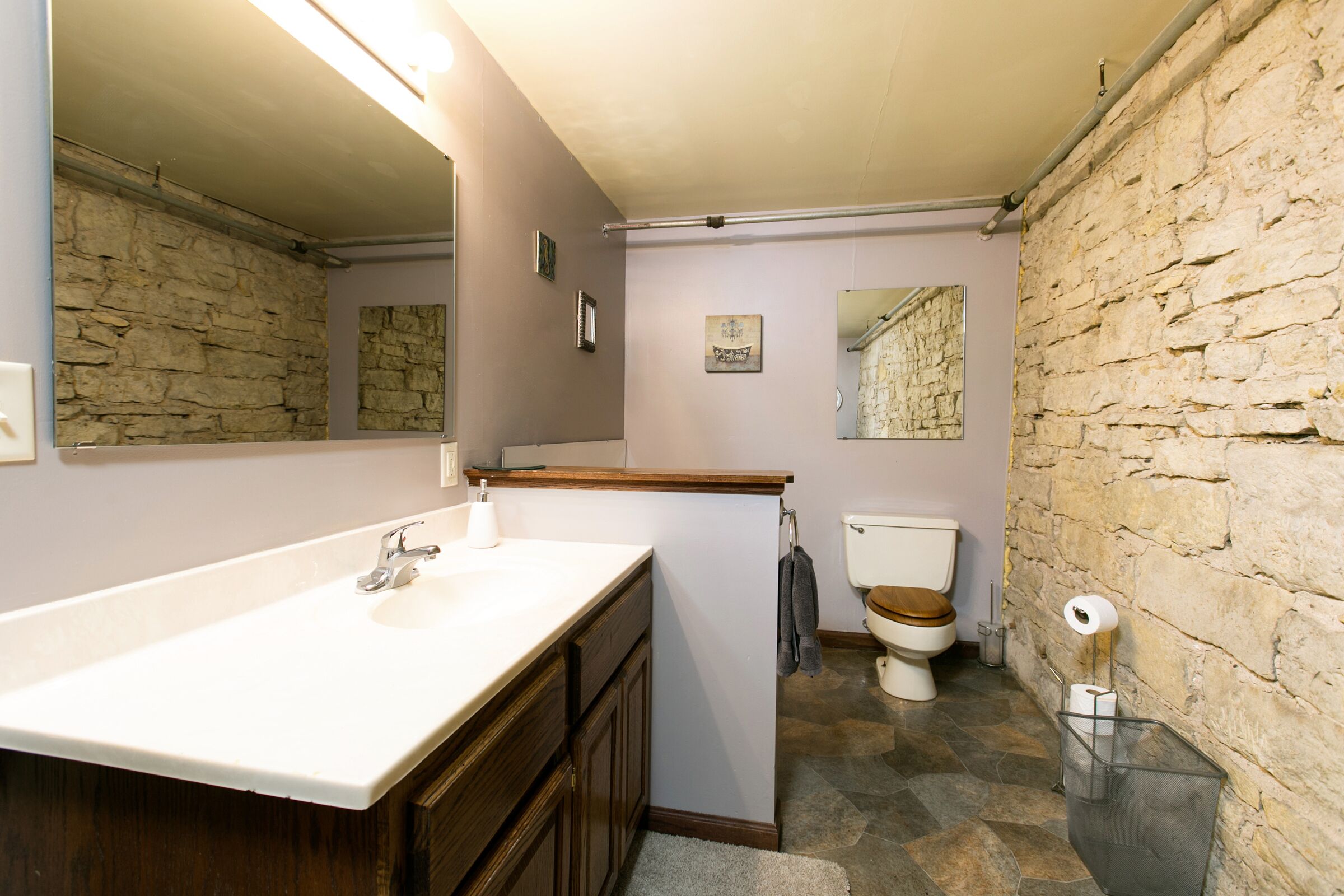 Un baño con pared de piedra, tocador de madera, lavabo blanco y un inodoro.